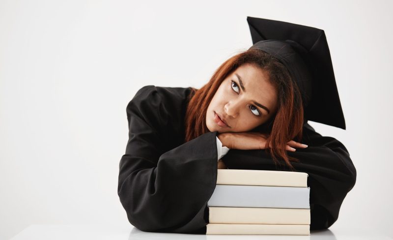 Upset beautiful african graduate lying on books thinking sitting over white background. Copy space.